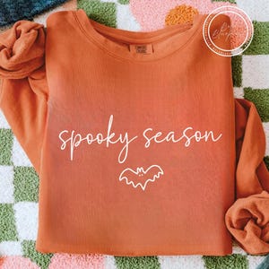 May include: An orange long-sleeved sweatshirt featuring the words "spooky season" in white script and a white bat illustration. The top is made of a soft material, ideal for the autumn season.