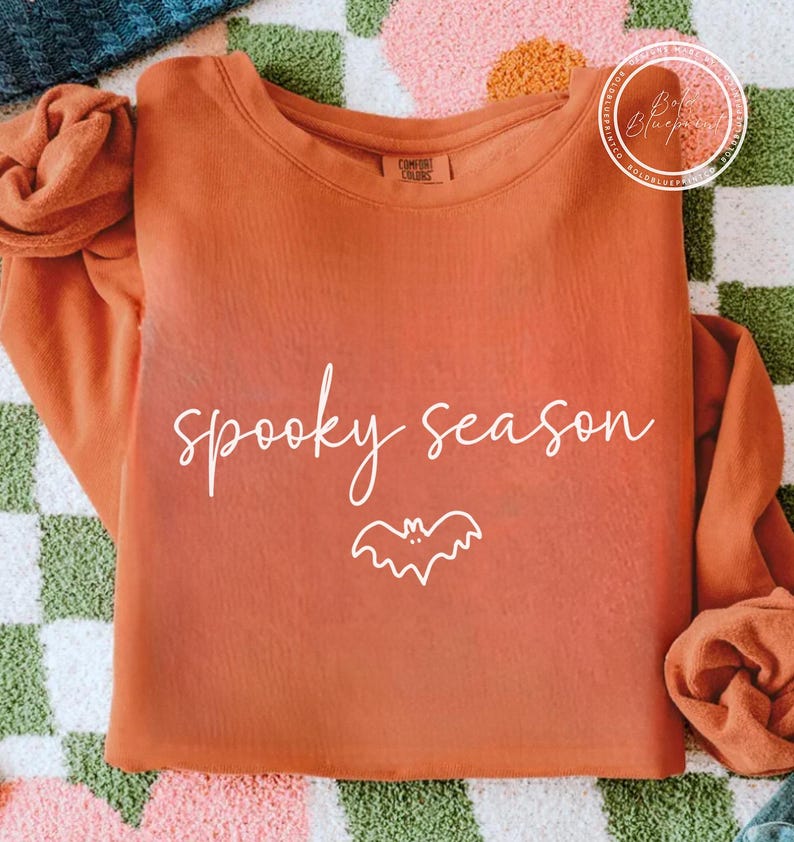 May include: An orange long-sleeved sweatshirt featuring the words "spooky season" in white script and a white bat illustration. The top is made of a soft material, ideal for the autumn season.