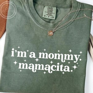 May include: Olive green t-shirt with the text "i'm a mommy. mamacita." in white, accented with starbursts. A gold necklace with a pendant rests on the shirt. The shirt has a Comfort Colors tag.