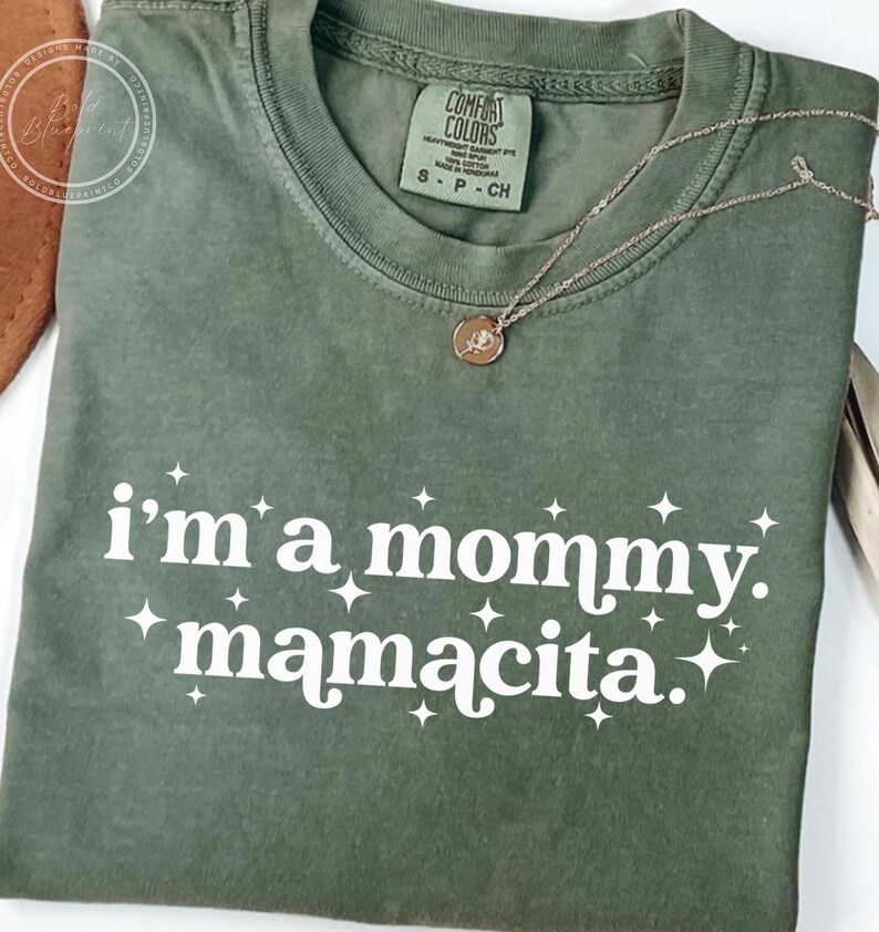 May include: Olive green t-shirt with the text "i'm a mommy. mamacita." in white, accented with starbursts. A gold necklace with a pendant rests on the shirt. The shirt has a Comfort Colors tag.