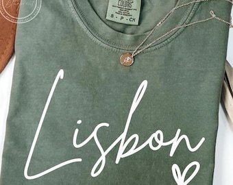 May include: Olive green t-shirt with the word "Lisbon" in white script and a small heart. A gold necklace with a pendant rests on the shirt. The shirt has a Comfort Colors label.
