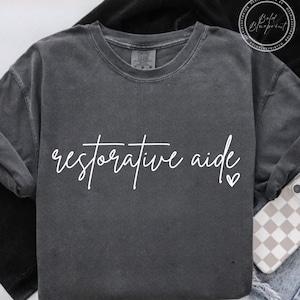 May include: A charcoal gray long-sleeved sweatshirt with the words "restorative aide" in white script. A small white heart is next to the word "aide". The sweatshirt is made of a soft material.