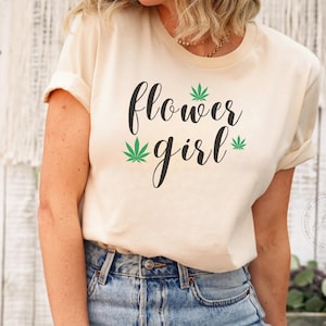 Op de afbeelding: Een wit T-shirt met de tekst "flower girl" in zwarte schrijfletters. Aan weerszijden van de tekst staan groene afbeeldingen van marihuanabladeren.