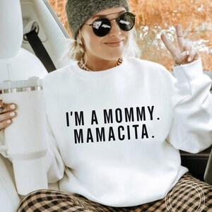 May include: White sweatshirt with the text "I'M A MOMMY. MAMACITA." in black. The person in the image is wearing a gray beanie, sunglasses, and a gold chain necklace. They are holding a white tumbler.