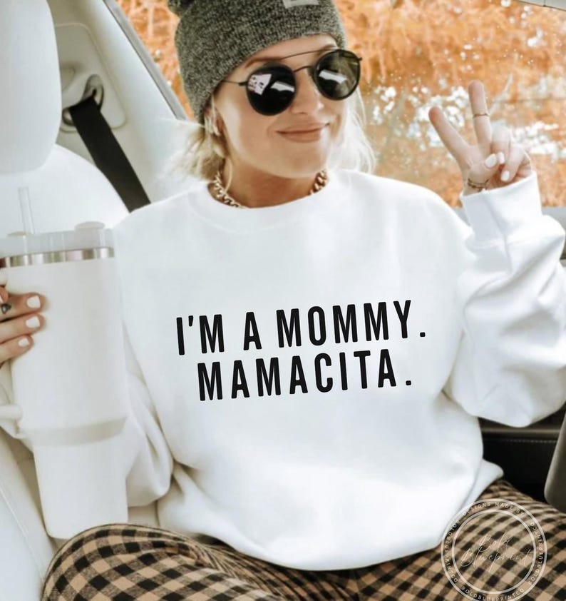 May include: White sweatshirt with the text "I'M A MOMMY. MAMACITA." in black. The person in the image is wearing a gray beanie, sunglasses, and a gold chain necklace. They are holding a white tumbler.