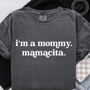 May include: Dark gray long-sleeve shirt with the text "i'm a mommy. mamacita." in white. The shirt has a crew neck and a relaxed fit. The shirt is made of a soft, comfortable material.