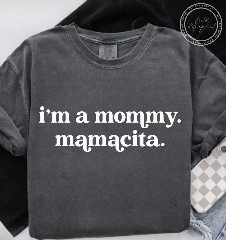 May include: Dark gray long-sleeve shirt with the text "i'm a mommy. mamacita." in white. The shirt has a crew neck and a relaxed fit. The shirt is made of a soft, comfortable material.