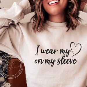 Op de afbeelding: Een crèmekleurig sweatshirt met de tekst "I wear my heart on my sleeve" in zwart gedrukt.