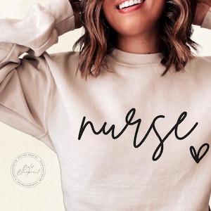 Peut inclure: Un sweat-shirt beige clair avec le mot "nurse" &eacute;crit en cursive noire et un petit c&oelig;ur noir &agrave; droite du mot.