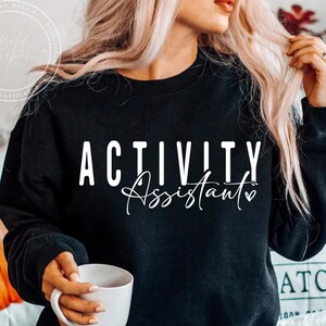 May include: Black crew neck sweatshirt featuring the text "ACTIVITY Assistant" in white, with a heart symbol. The image shows a white mug. The sweatshirt is a casual style.