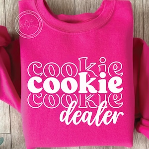 Könnte beinhalten: Ein leuchtend pinkfarbenes Sweatshirt mit dem weißen Schriftzug "Cookie Dealer", der vertikal angeordnet ist. Der Text hat eine verspielte, umrandete Schriftart. Das Sweatshirt hat einen Rundhalsausschnitt und lange Ärmel.