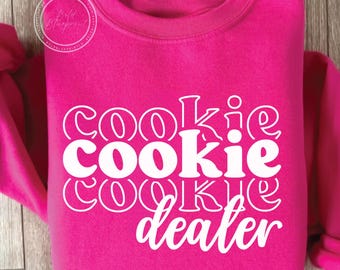 May include: A bright pink sweatshirt with the words "Cookie Dealer" in white, stacked vertically. The text has a playful, outlined font. The sweatshirt has a crew neck and long sleeves.