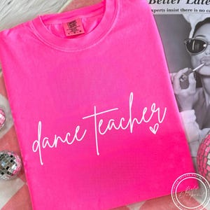 Peut inclure: Un t-shirt rose vif avec l'inscription "dance teacher" en écriture blanche et un petit cœur. Le t-shirt est plié, suggérant un tissu doux. L'ensemble est décontracté et lumineux.