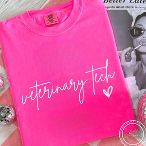 May include: Bright pink t-shirt with the words "Veterinary Tech" in white script and a small heart. The shirt is folded and lies on a pink and white checkered surface. The shirt has a crew neck and short sleeves.