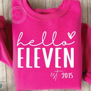 Könnte beinhalten: Ein leuchtend pinkfarbenes Sweatshirt mit dem weißen Schriftzug "hello ELEVEN", einem Herz und "est. 2015" darunter. Das Sweatshirt hat einen Rundhalsausschnitt.
