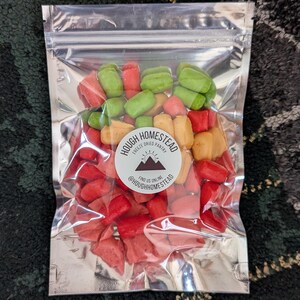 May include: A transparent, sealed bag containing a variety of freeze-dried sweets in red, green, and yellow. The bag features a circular sticker with the text "Hough Homestead Freeze Dried Pantry" and the Instagram handle @houghhomestead.