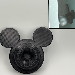 May include: Black 3D-printed Mickey Mouse-shaped door knob cover. The cover has a textured surface and is designed to fit over a standard doorknob. The design includes two circular ears and a circular base.