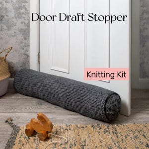 May include: A gray knitted door draft stopper resting against a white door. The text "Door Draft Stopper" is above the stopper, and "Knitting Kit" is in a pink box. A wooden pull-along toy and a woven basket are in the background.