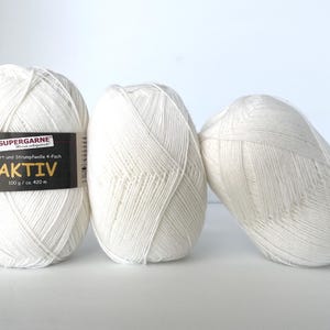 May include: Three off-white yarn skeins. One skein has a black and white label that reads "SUPERGARNE AKTIV" and "100 g / ca. 420 m". The yarn is tightly wound and appears soft. The skeins are arranged on a white surface.
