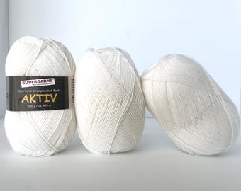 Puede incluir: Tres ovillos de hilo blanco roto. Uno de ellos tiene una etiqueta en blanco y negro que dice "SUPERGARNE AKTIV" y "100 g / ca. 420 m". El hilo está bien enrollado y parece suave. Los ovillos están colocados sobre una superficie blanca.