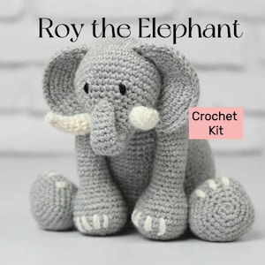 May include: A crocheted gray elephant toy with white tusks and accents. The elephant has large ears and is sitting down. The text "Roy the Elephant" is above the elephant, and "Crochet Kit" is in a pink box.
