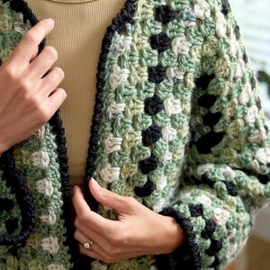 May include: A crochet cardigan featuring a granny square pattern in shades of sage green, cream, and black. The garment has a black crocheted border along the front opening and cuffs, constructed with a textured yarn in a classic open-stitch design.