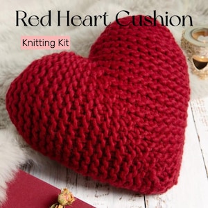 May include: A knitted red heart-shaped cushion. The cushion is made from a thick, textured yarn and is displayed on a white wooden surface. The text "Red Heart Cushion" and "Knitting Kit" are visible.