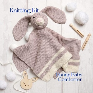 May include: A knitted bunny baby comforter in soft lavender and cream. The comforter has a bunny head and a blanket with cream stripes and embroidered details. The image also includes wooden clothespins, white pom-poms, and a wooden bunny cutout. Text reads "Knitting Kit" and "Bunny Baby Comforter."
