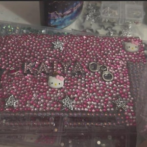 May include: A pink license plate frame and matching phone case, both heavily embellished with small, sparkling rhinestones. The license plate frame features the name "KAYA" and two Hello Kitty faces. Silver star and handcuff charms add detail.