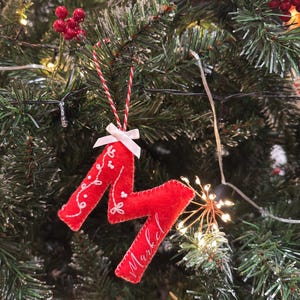 Könnte beinhalten: Roter Weihnachtsbaumschmuck aus Filz in Form des Buchstabens "M" mit dem Namen "Markel" in weißer Schrift. Der Schmuck ist mit weißer floraler Stickerei und einer kleinen weißen Schleife verziert und wird an einer rot-weißen Schnur aufgehängt.