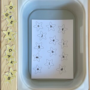 May include: A white tray with a sheet of paper inside. The paper has a grid of yellow stars with letters printed inside each star. The letters are: c, h, f, j, e, l, g, b, k, m, i, a, d.  There are also yellow stars with letters on the side of the tray. The letters are: o, g, e, f, s, c, a, k, b.