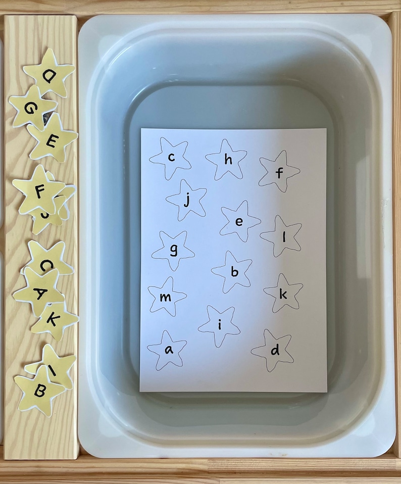 May include: A white tray with a sheet of paper inside. The paper has a grid of yellow stars with letters printed inside each star. The letters are: c, h, f, j, e, l, g, b, k, m, i, a, d.  There are also yellow stars with letters on the side of the tray. The letters are: o, g, e, f, s, c, a, k, b.