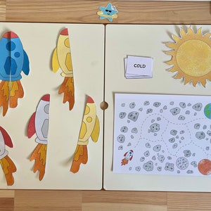 May include: A set of educational materials for children featuring a yellow sun, a blue rocket, a white rocket, a yellow rocket, a white card with the word "COLD" printed on it, and a black and white illustration of a space scene with a rocket, a planet, and asteroids.