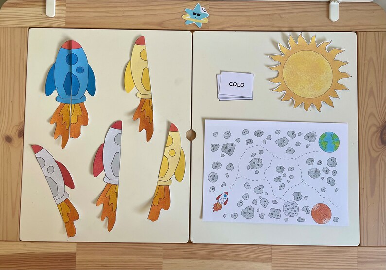 May include: A set of educational materials for children featuring a yellow sun, a blue rocket, a white rocket, a yellow rocket, a white card with the word "COLD" printed on it, and a black and white illustration of a space scene with a rocket, a planet, and asteroids.