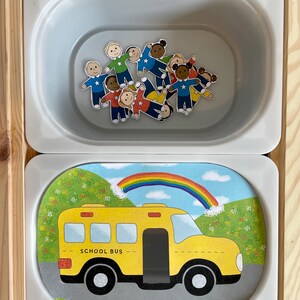 May include: A white oval container with a yellow school bus with a rainbow above it. The bus has the words "SCHOOL BUS" on the side. There are also small paper dolls inside the container.