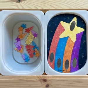 May include: A white plastic bin filled with colorful star-shaped pieces. Next to the bin is a white plastic panel with a rainbow-colored shooting star design and the text "Bright Little Beings".