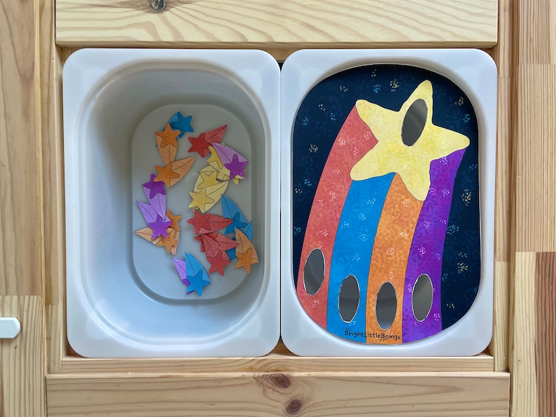 May include: A white plastic bin filled with colorful star-shaped pieces. Next to the bin is a white plastic panel with a rainbow-colored shooting star design and the text "Bright Little Beings".