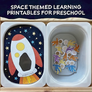 May include: A white tray with a rocket ship illustration on a black background with white stars. The rocket ship has a yellow flame and a black window. A second white tray contains a collection of astronaut figures.
