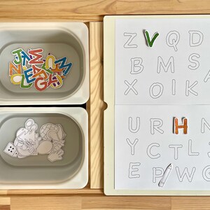 May include: A white board with a grid of letters, some of which have colorful letters placed on top.  The letters on the board are: Z, V, Q, D, B, M, S, A, X, O, I, K, U, R, H, N, Y, C, T, L, J, E, P, W, G.  There is a flower and a pencil drawn on the board.  Two white containers with lids are below the board.  One container has colorful letters inside and the other container has a drawing of a person with a globe and a dice.