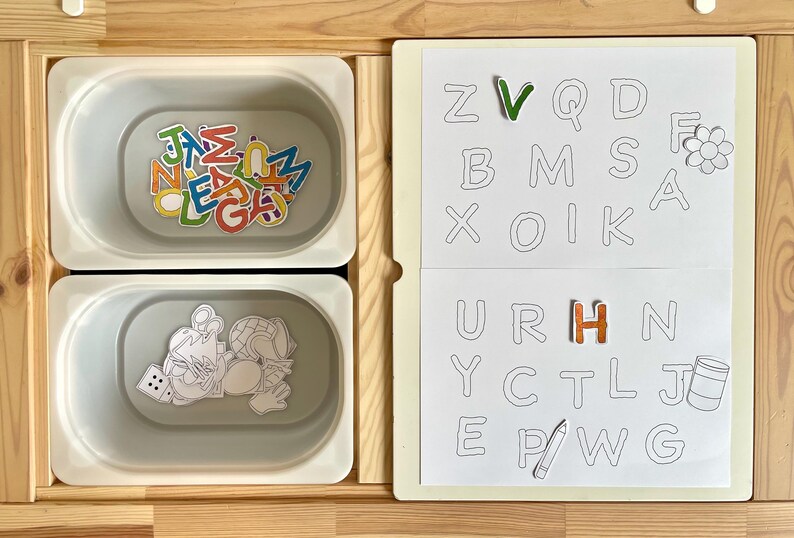 May include: A white board with a grid of letters, some of which have colorful letters placed on top.  The letters on the board are: Z, V, Q, D, B, M, S, A, X, O, I, K, U, R, H, N, Y, C, T, L, J, E, P, W, G.  There is a flower and a pencil drawn on the board.  Two white containers with lids are below the board.  One container has colorful letters inside and the other container has a drawing of a person with a globe and a dice.