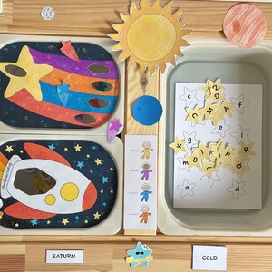 May include: A wooden playset with a space theme. The set includes a rocket, a sun, planets, stars, and astronauts. There is a container with letter tiles in the shape of stars. The words "Saturn" and "Cold" are written on the wooden surface.