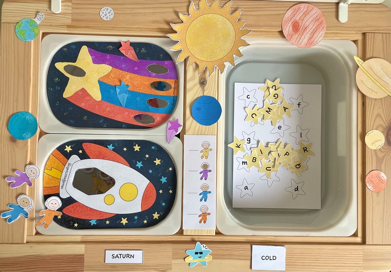 May include: A wooden playset with a space theme. The set includes a rocket, a sun, planets, stars, and astronauts. There is a container with letter tiles in the shape of stars. The words "Saturn" and "Cold" are written on the wooden surface.