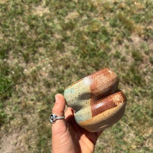 Puede incluir: Una pequeña taza de cerámica hecha a mano con un cuerpo redondeado y con hoyuelos. El exterior presenta un esmalte moteado con una paleta de colores de marrón tierra, beige suave y verde salvia, mostrando una clara división horizontal.