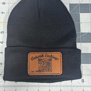 May include: A black beanie with a brown rectangular patch. The patch features the text "Artistik Etchings" and a QR code. The text "est. '24" is also visible on the patch. The beanie is made of a knitted material.