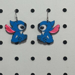 May include: A pair of blue and pink cartoon character earrings. The earrings feature a blue character with large eyes and pink ears. The earrings are hanging from silver hooks. The background is a white surface with black dots.