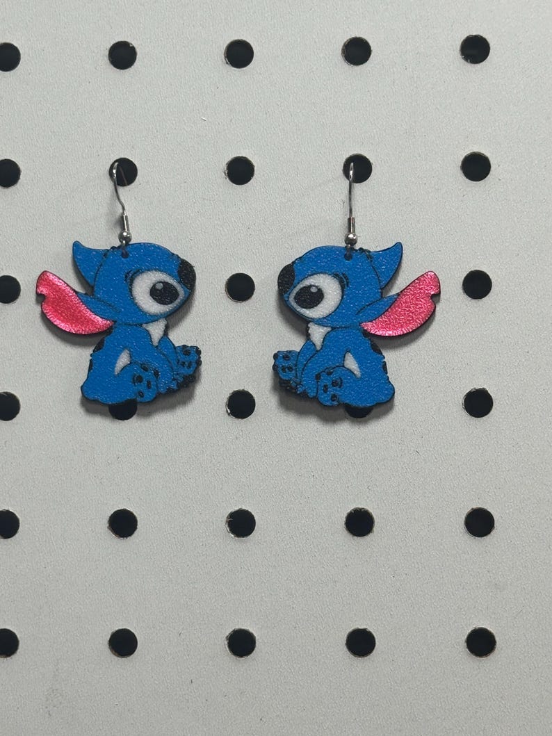May include: A pair of blue and pink cartoon character earrings. The earrings feature a blue character with large eyes and pink ears. The earrings are hanging from silver hooks. The background is a white surface with black dots.