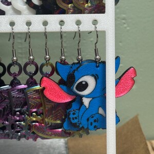 May include: A display of handmade earrings. One pair features a blue cartoon character with pink ears. Other earrings include colorful metal designs and circular shapes. The earrings hang from a white display rack.