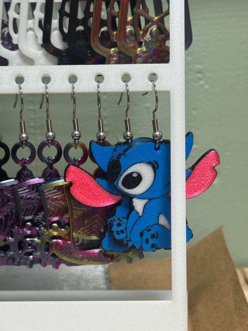 May include: A display of handmade earrings. One pair features a blue cartoon character with pink ears. Other earrings include colorful metal designs and circular shapes. The earrings hang from a white display rack.