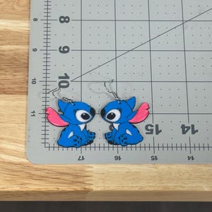 May include: A pair of blue Stitch character earrings with pink ears and large eyes. The earrings are on a gray cutting mat with inch measurements. The earrings are made of a shiny material and have silver hooks.