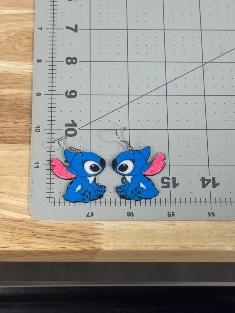 May include: A pair of blue Stitch character earrings with pink ears and large eyes. The earrings are on a gray cutting mat with inch measurements. The earrings are made of a shiny material and have silver hooks.
